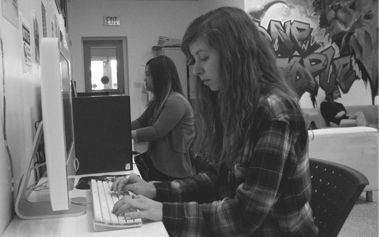 Two young women sit beside each other on their computers, while one of them gazes solemnly at her keyboard. 