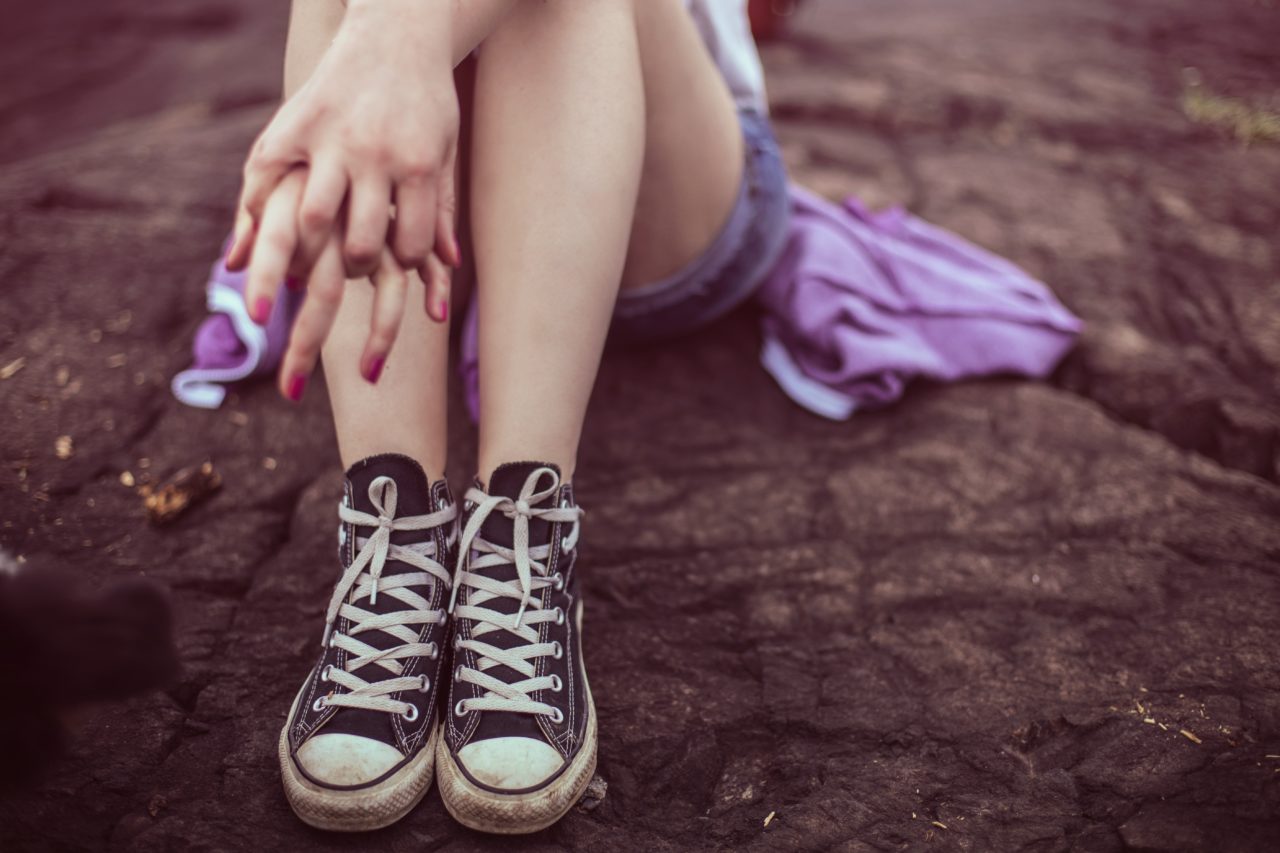Young woman pictures sitting on rocky ground, wearing converse sneakers and photo is cropped below her knees. 