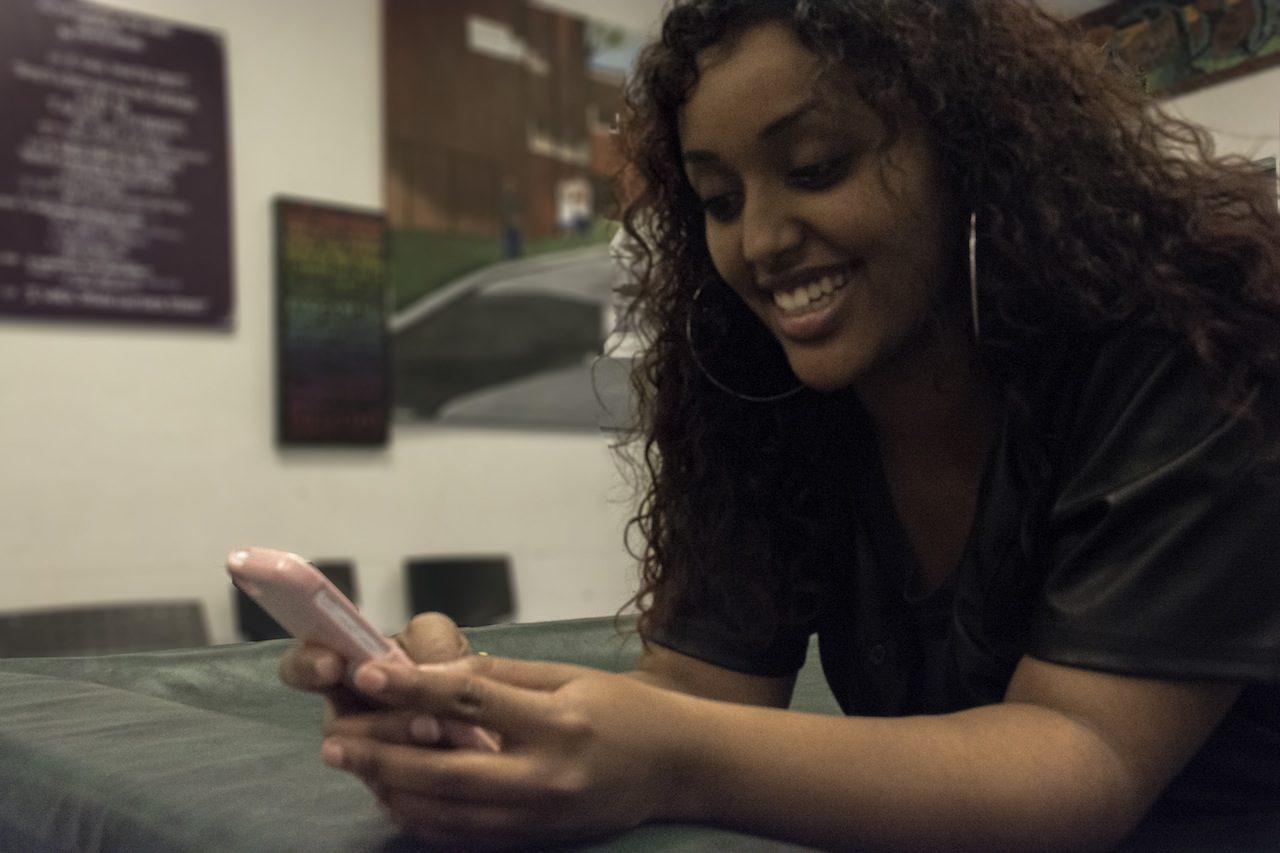 Young racialized woman looking at her phone screen and smiling, while laying on her stomach. 