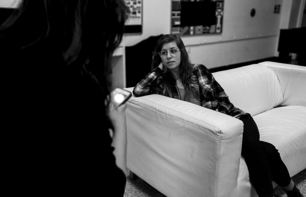 Young woman resting head on her arm and sitting on white couch looks to her right at a friend.