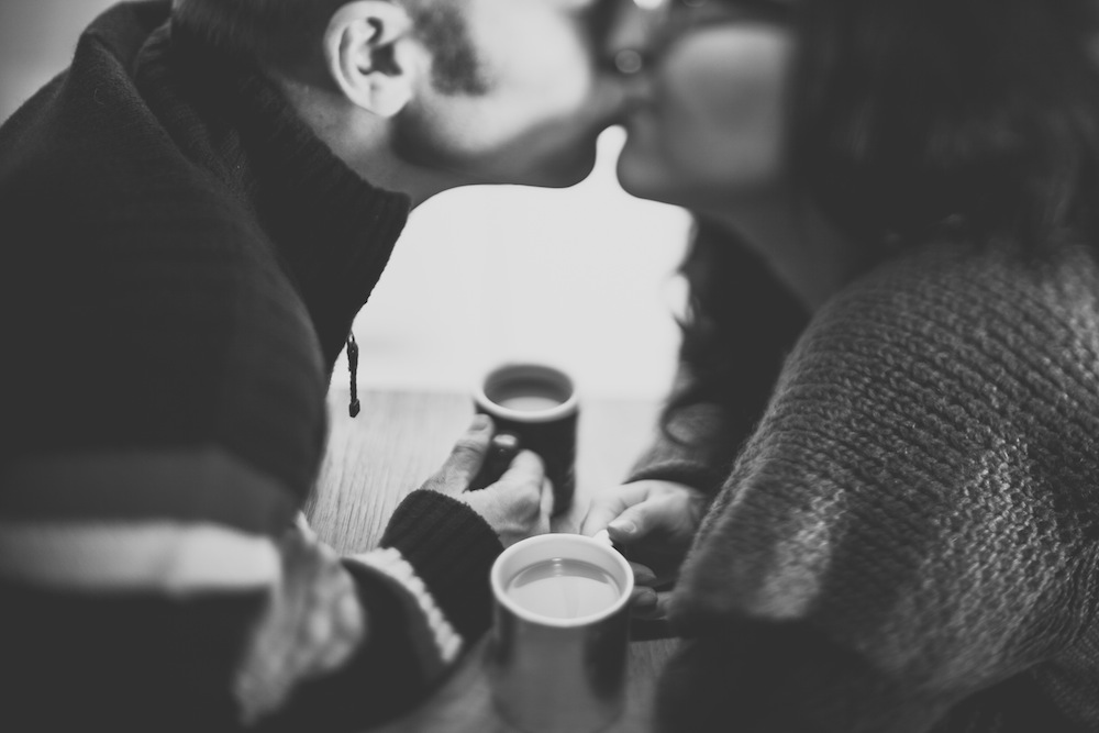 A couple kisses over mugs of coffee. 