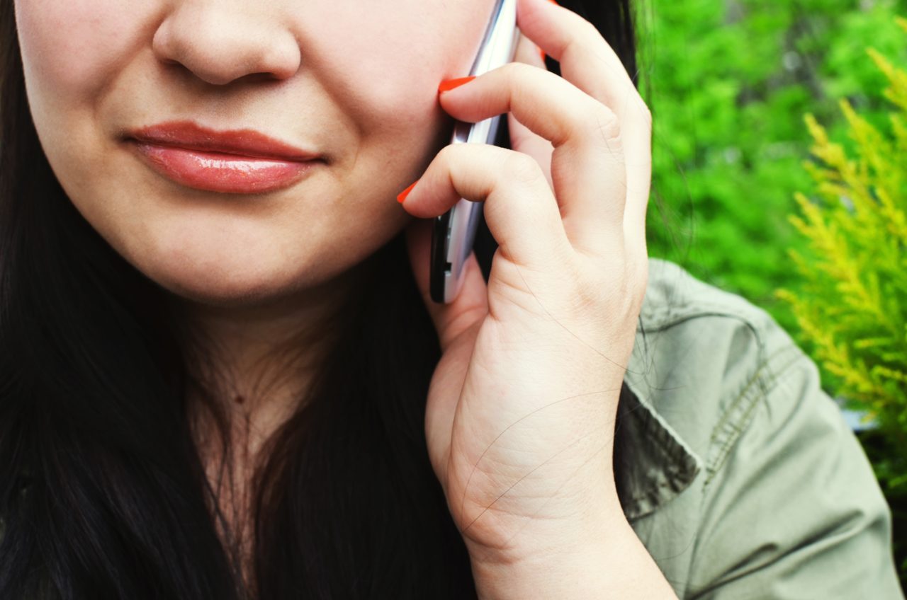 Young woman on phone, image is cropped at her nose, mouth and hand holding phone with a green leafy bush-like background. 