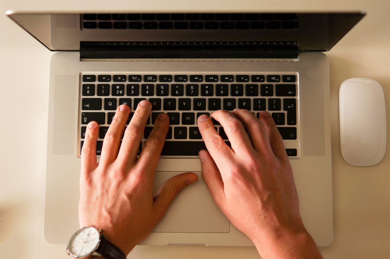 A laptop depicted in birds eye view, with a pair of men's hands at the keyboard. 